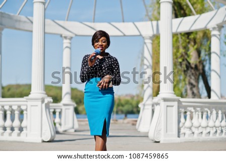 Stylish african american model girl TV presenter with microphone in glasses, blue skirt and black blouse posed outdoor against white stone arch with mobile phone.