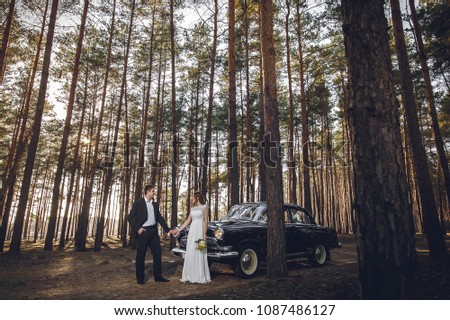 Just married wedding couple is standing near the retro vintage car in the park. Spring or autumn sunny day in forest. bride in elegant white dress with bouquet and elegant groom in love hugging.