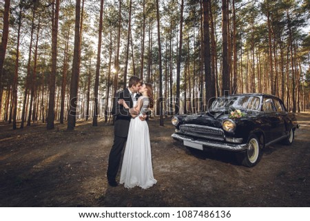 Just married wedding couple is standing near the retro vintage car in the park. Spring or autumn sunny day in forest. bride in elegant white dress with bouquet and elegant groom in love hugging.