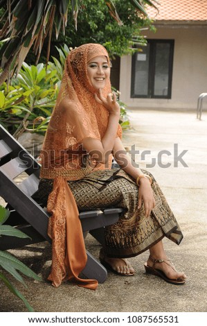 Woman in Thai tradition Muslim dress in brown color sitting on the pool chair beside the pool and roof background.