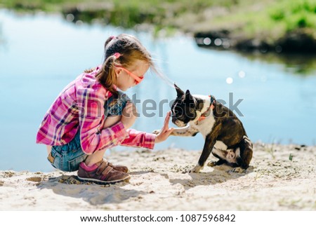 Child girl playing with boston terrier dog on the sandy river bank outdoors