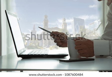 A man holding a mug with steaming coffee, pointing with a pen on the laptop screen on the background of a window with a panorama of the city
