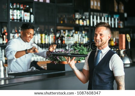 Smiling elegant waiter is holding a tray with a decorated cocktail with a smiling mixed race male expert bartender at the bar counter at the background Royalty-Free Stock Photo #1087608347