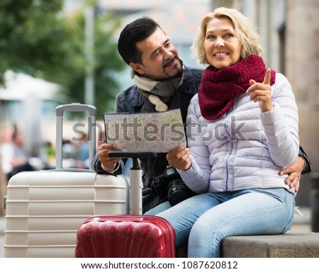 Happy smiling mature couple with suitcases and camera checking direction with map
