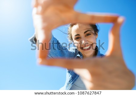 portrait of a young smiling attractive woman in jeans clothes at sunny day on the blue sky background. woman shows a frame from hands like photo. Photo Frame Hands Made By A Hipster young girl