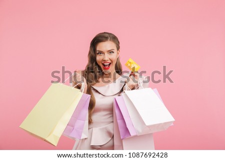 Photo of rich fashion woman 20s in dress smiling while holding different shopping bags and credit card in hands isolated over pink background