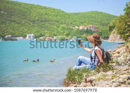 girl traveler sits on a mountain lake and takes pictures Girl takes photo for travel blog. View from back of the tourist traveler