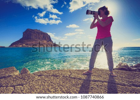 Woman with camera taking photo on seacoast. Rocky island Monemvasia in the background. Greece, Peloponnese, Lakonia. Travel destinations.