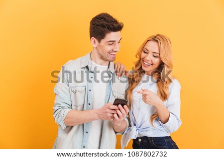 Photo of happy people man and woman laughing while pointing finger at smartphone isolated over yellow background Royalty-Free Stock Photo #1087802732