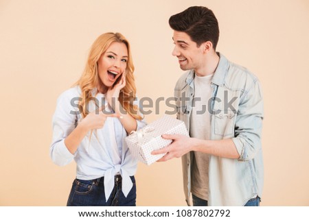 Image of beautiful young woman pointing finger at birthday box holding by attractive brunette man isolated over beige background