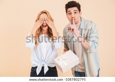 Image of brunette man with present box keeping finger at lips and showing shh while standing near woman covering eyes with hands isolated over beige background