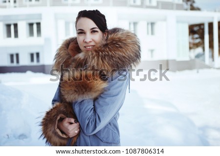 Beautiful brunette girl with two pigtails in a blue coat with a brown fluffy fur collar in the winter on nature. Snowy spruce on the background.
