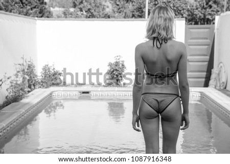 Back view of unrecognizable young woman in bikini standing on poolside on black and white. 