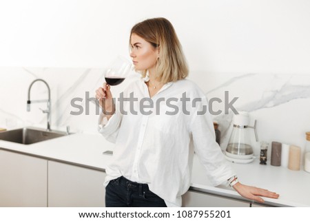 Portrait of young lady standing with glass of red wine in hand and dreamily looking aside at home isolated