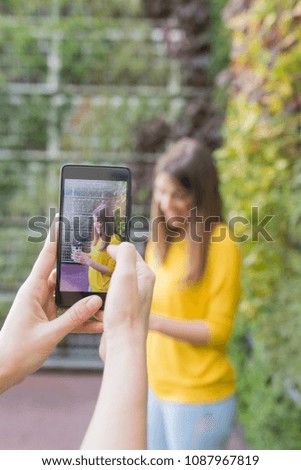 two beautiful women taking selfie with mobile phone. green background. One is holding a cup of coffee. They are laughing . Outdoors lifestyle and friendship