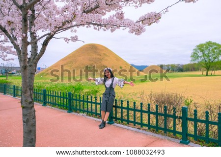 Young woman sitting on the fence with view of tomb and cherry blossom durign Spring season at Gyeongju city of South Korea.