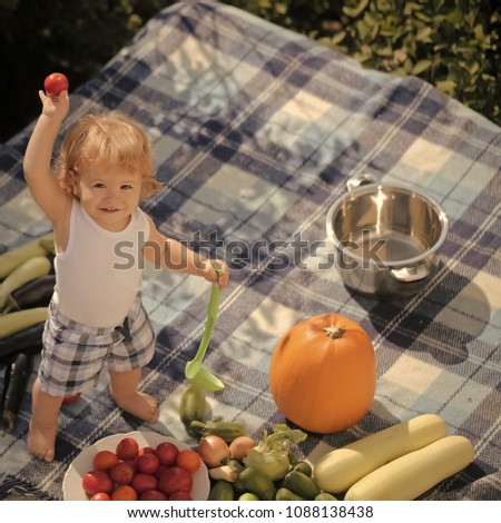 One little cook smiling boy at picnic standing with ladle pot orange pumpkin squash and cucumber holding red tomato with food sitting on checkered plaid on natural background sunny day, square picture