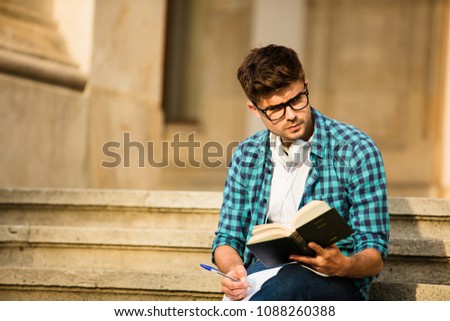 young student with glasses standing on stairs of school or university, looking over his homework, reading a book and taking notes