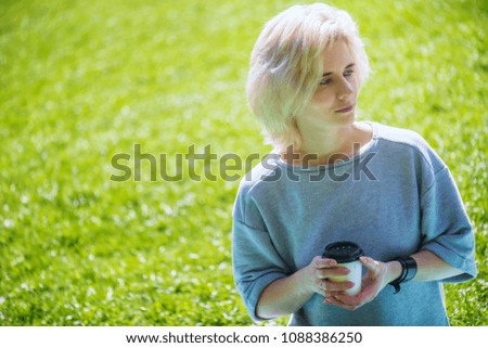 Young beautiful blonde woman sitting in the summer park with paper cup of coffee. 