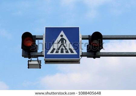 Pedestrians crossing blue and white road sign with two blinking yellow lights mounted on metal frame on cloudy sky background