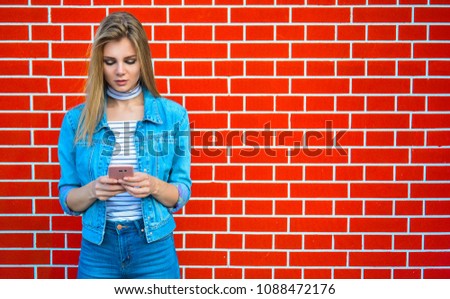 Young pretty woman type message - Female jeans fashion portrait - Teenager student holding mobile phone for sms next to brick wall background