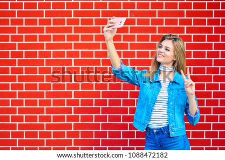 Young pretty woman taking selfie outdoors - Female summer fashion portrait - Teenager student holding mobile phone for selfi photo next to brick wall background