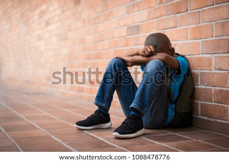 Young boy sitting alone with sad feeling at school. Depressed african child abandoned in a corridor and leaning against brick wall. Bullying, discrimination and racism concept with copy space. Royalty-Free Stock Photo #1088478776