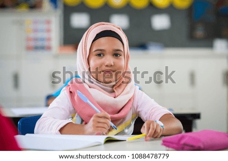 Happy young smiling girl wearing hijab and looking at camera while sitting at desk with exercise book Elementary muslim schoolgirl writing notes in classroom. Portrait of arab school girl in chador.