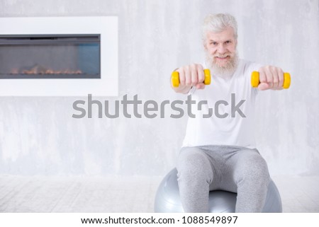 Exercising stamina. Pleasant senior man holding dumbbells and exercising the strength of his arms while sitting on the fitness ball