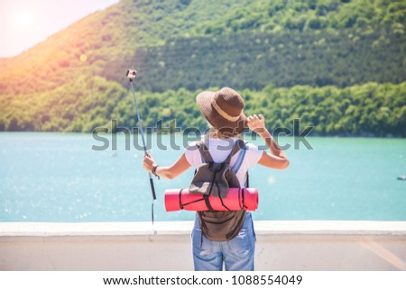 girl traveler makes selfi with an action camera on a mountain lake. She makes a photo for travel blog. View from back of the tourist traveler