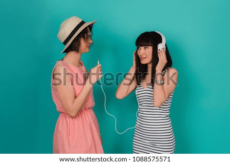 Two cheerful young women with earphones listening to music and talking selfie over pink background