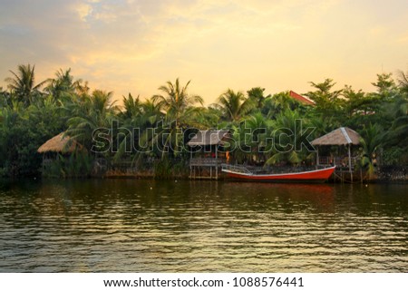 Pristine riverbank at sunset, palms trees, a red boat and bungalows, situated at the Praek Tuek Chhu River in Kampot Province in southern Cambodia, Asia. Natural asian riverside landscape Royalty-Free Stock Photo #1088576441