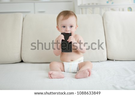 Adorable toddler boy sitting on sofa and biting smartphone