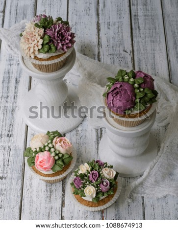 
Cupcakes decorated with cream flowers: roses, peonies, chrysanthemums, poppies, scabiosa, on a wooden background with a lacy napkin