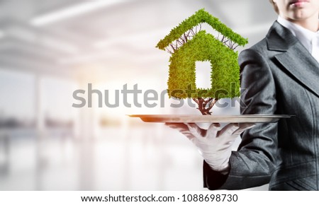 Closeup of waitress's hand in white glove presenting green plant in form of house sign on metal tray with office view on background.