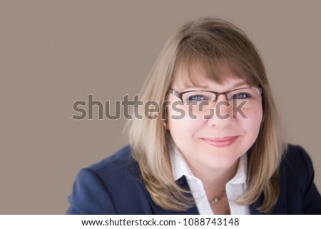 Head shot of professional business woman in her upper forties with blonde hair and blue eyes wearing a blue suit jacket, white blouse, necklace, earrings and glasses. Horizontal landscape orientation.