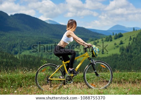 woman cyclist riding on yellow bicycle on a rural trail in the mountains, enjoying valley view on sunny day. Mountains, forests and blue sky on the blurred background. Outdoor sport activity