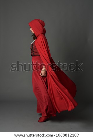 full length portrait of brunette girl wearing red fantasy costume with cloak, standing pose on a guy studio background.