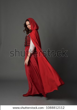full length portrait of brunette girl wearing red fantasy costume with cloak, standing pose on a guy studio background. Royalty-Free Stock Photo #1088812112