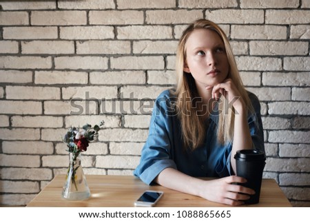 Young blonde in blue shirt sits at a table in a cafe on which stands a black paper cup with coffee and lies a phone on a brick wall background. The teen looks up thoughtfully and holds in her hand