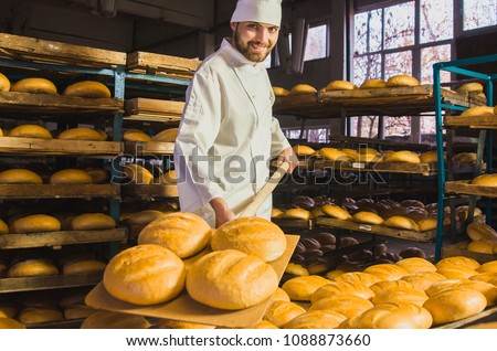 Baker. A young handsome bakery worker on the background of bread, takes bread from a stove with a wooden shovel. Industrial production of bakery products. a man in the baker's special clothing. Bakery Royalty-Free Stock Photo #1088873660
