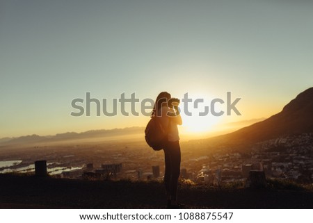 Woman with camera taking photo of a landscape. Female traveler shooting pictures from a mountain top at sunset.