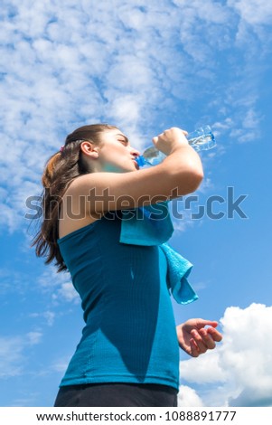 Beautifull girl runner is having break, drinking water against clear blue sky background.