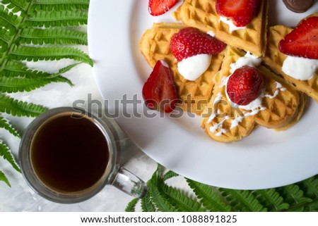 Waffles with strawberry, a cup of coffee and green leaves of fern on a table. Beautiful and tasty breakfast.