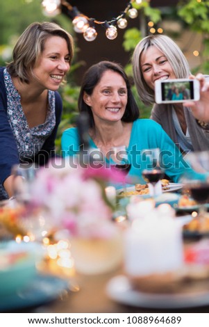 Summertime. Group of friends gathered around a table in the garden to share a meal. Three beautiful women in their forties taking a selfie