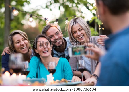 Some friends in their forties gathered around a table in the garden for a good time. A man takes a picture of a group of friends