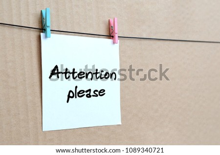 Attention please. Note is written on a white sticker that hangs with a clothespin on a rope on a background of brown cardboard