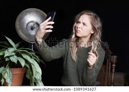 Unconfident student lady with blonde wavy hair taking pictures with smart phone, making faces at the camera. Young office female manager in bad mood making selfie using electronic device indoor.