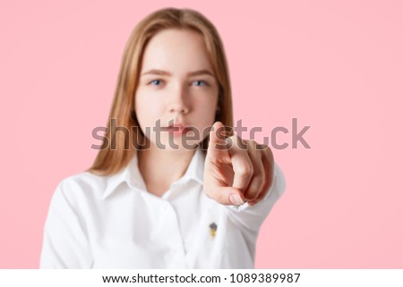 Lovely serious confident young female points at camera with fore figer, chooses someone, isolated on pink background, focus on hand, blurred background. I choose you! People and choice concept