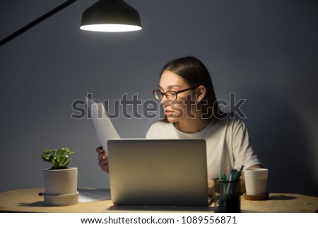 Female student getting ready for exam, reading paper handout material, working at laptop, or preparing presentation at night under lamp light. Woman taking online business course in small home office